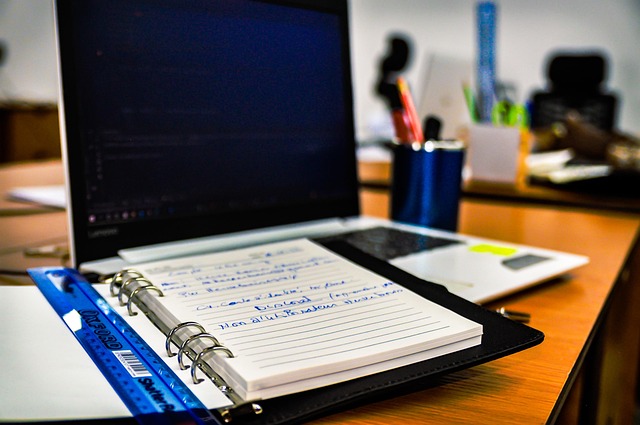 ノートパソコンとリングノートが並ぶデスク。手書きのメモとデジタル記録を一元管理するワークスペースを表現。