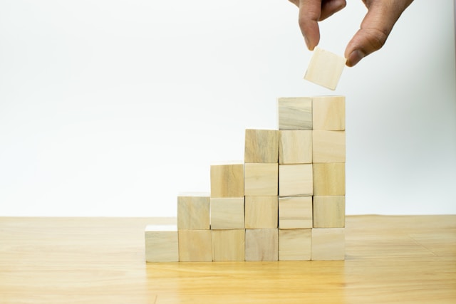 木製のブロックを階段状に積み重ね、その上にさらにブロックを置こうとする手。日報を継続することで一歩一歩成長を重ねる様子を象徴している。