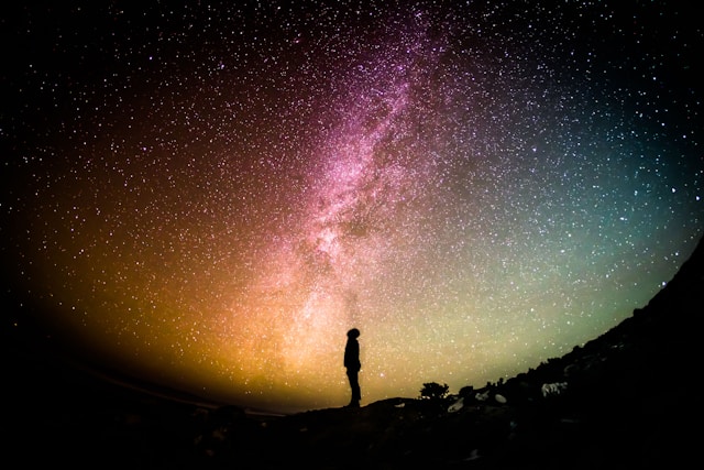 広大な星空と天の川を背景にシルエットの人物が立っており、色とりどりの銀河が幻想的に広がる風景