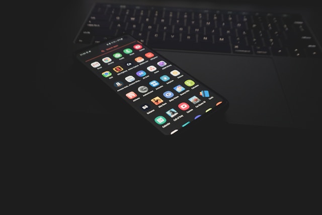 暗いデスクの上にノートパソコンとアプリが並んだスマートフォンが置かれている。営業日報アプリを含むモバイルアプリの選び方や機能比較を想起させるイメージ。