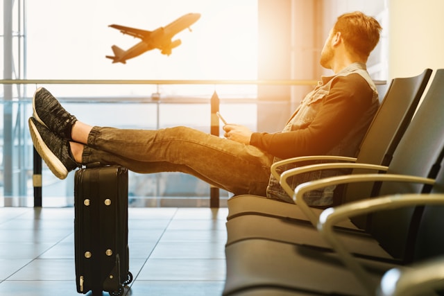 空港のロビーでスーツケースに足を乗せ、スマホを手に飛行機を眺める男性の写真。旅行中に翻訳アプリを使うシーンを表現しています。
