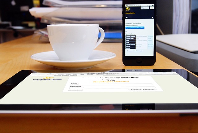 デスクにタブレットとスマートフォン、白いコーヒーカップが並び、タブレットにはウェブサイトが表示されている。オンラインツールを使ってマニュアルを作成・編集しているような現代的な作業環境を表す。
